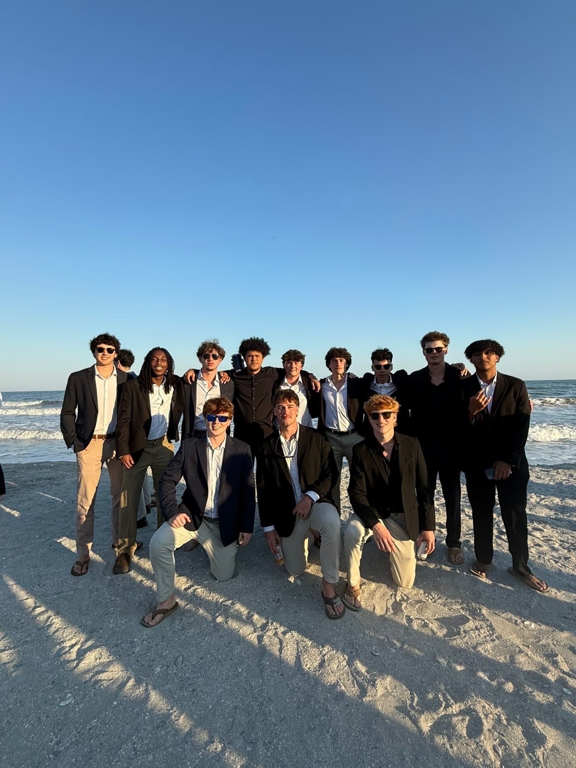 A large group of brothers in suits and blazers posed together on the beach