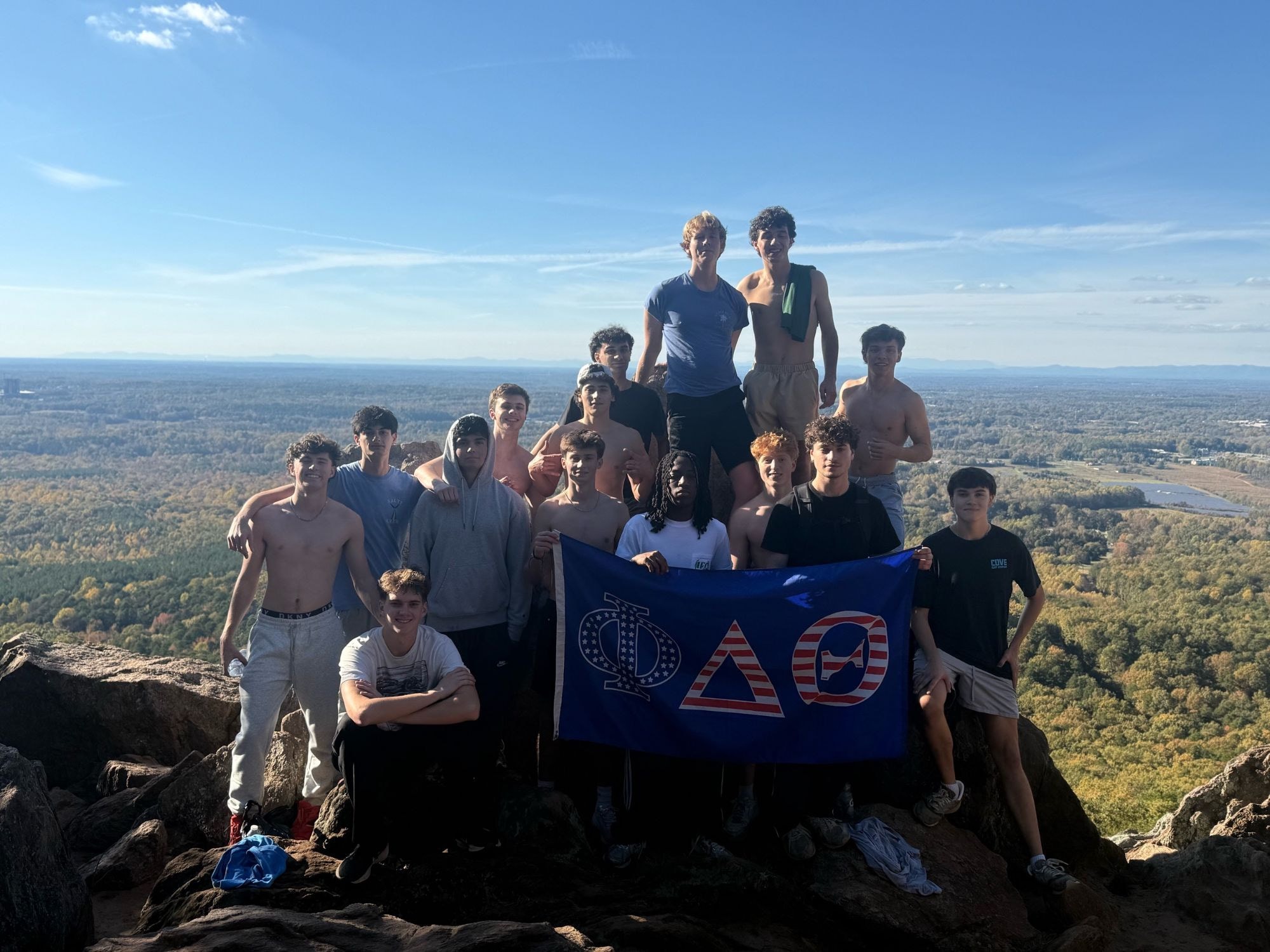 Brothers on a rocky summit holding a red, white and blue Phi Delta Theta flag, with autumn forest stretching to distant mountains